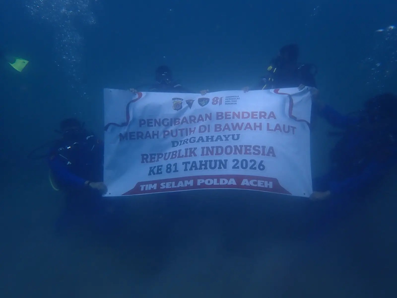 Bendera Merah Putih tampak terbentang di antara terumbu karang dasar laut Krueng Raya, menjadi simbol kedaulatan Indonesia sebagai negara maritim.